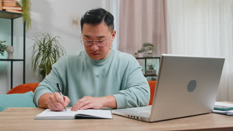 Asian-man-freelancer-making-notes-on-notebook-at-home,-use-laptop,-prepare-business-financial-report