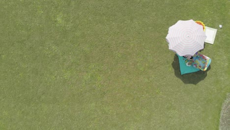 Looking-down-at-a-mother-and-child-enjoying-a-warm-day-under-an-umbrella-playing-with-toys,-aerial-reveal