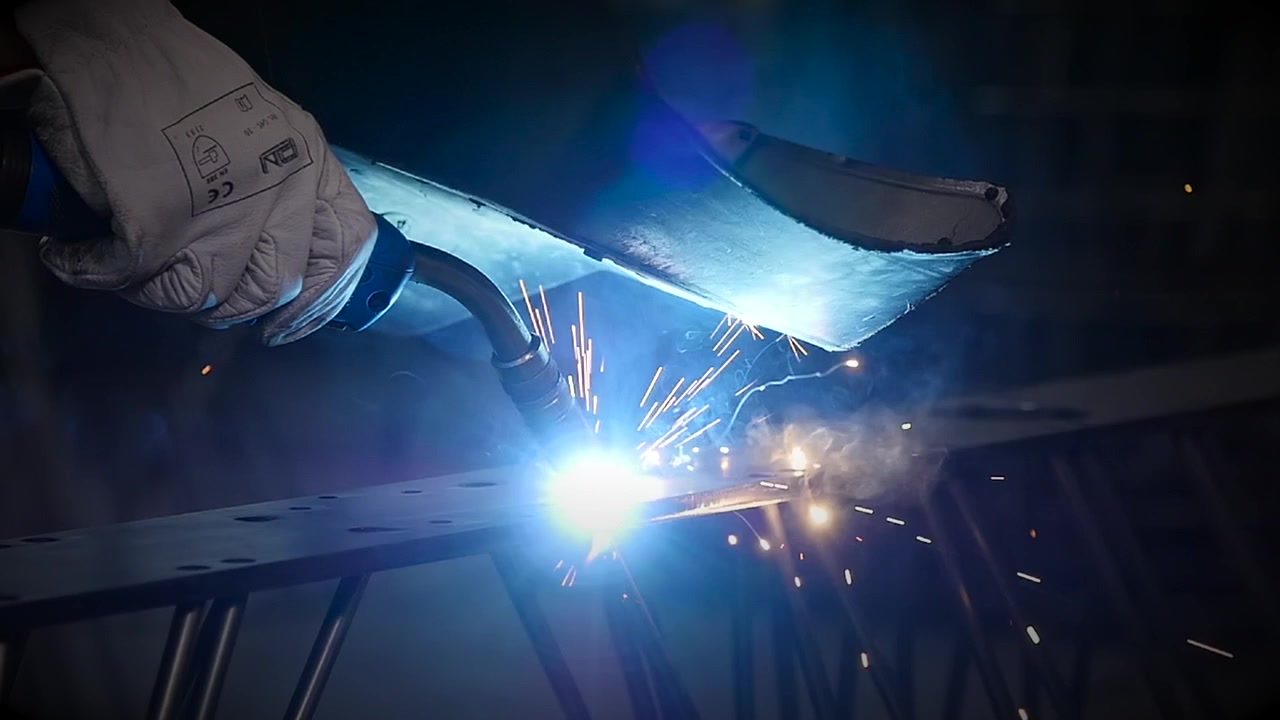 Slow-motion close-up of MIG welding on a steel plate with bright electric arc, sparks and smoke inside an industrial workshop.