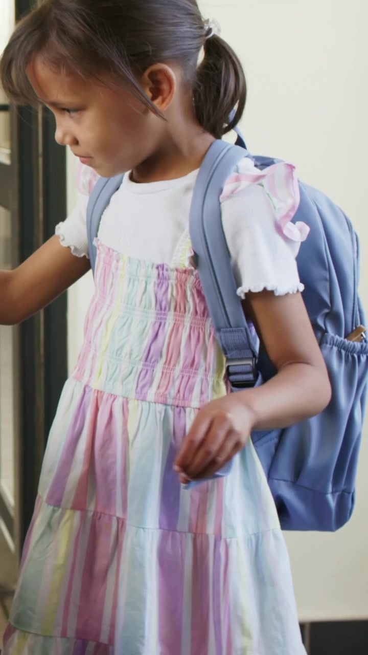 Vertical video: Adjusting light blue backpack, girl preparing for school in foyer, smoothing dress