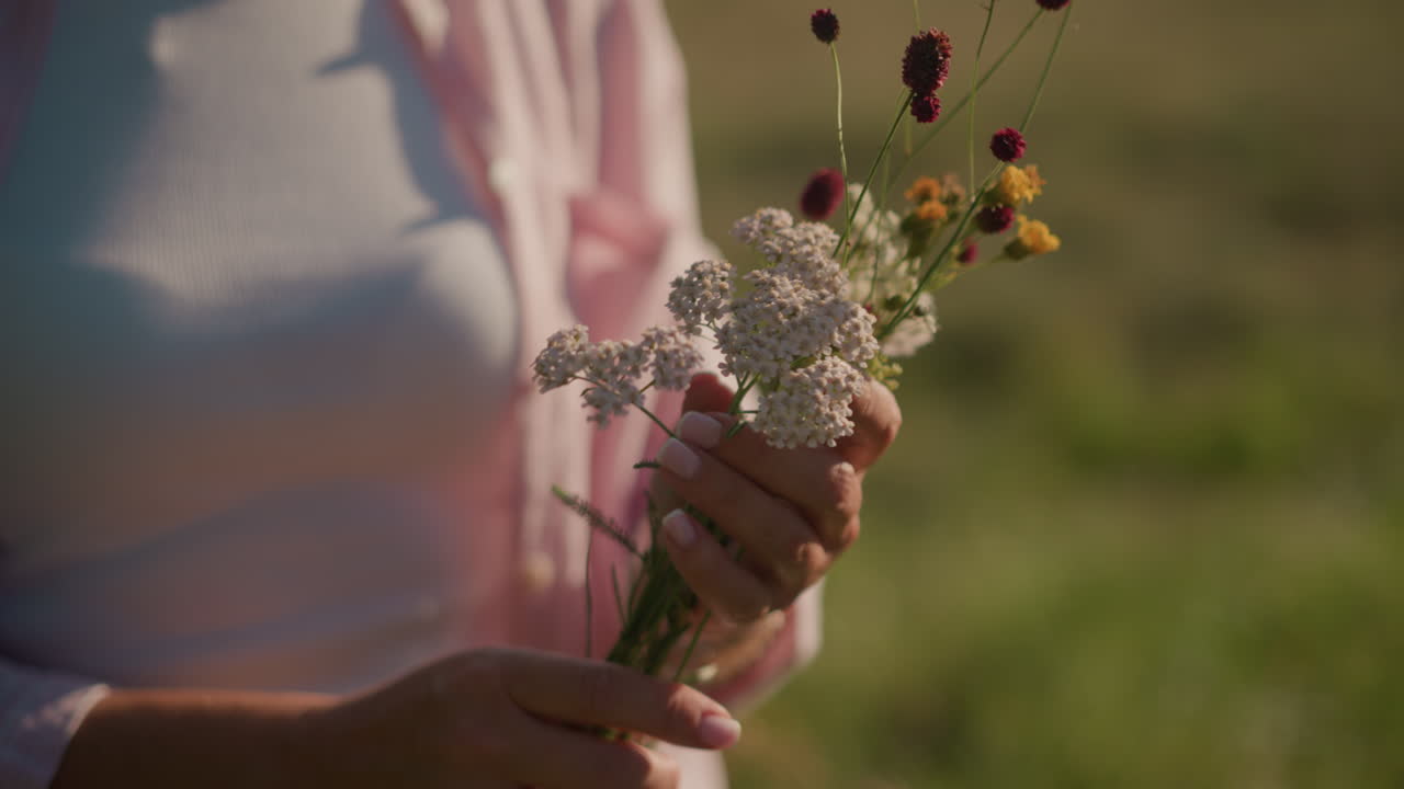 primer plano de un jardinero con una camisa rosa abierta y un polo blanco, arreglando cuidadosamente las flores en las manos, con vibrantes flores rojas, amarillas y blancas contra el fondo de un campo de hierba