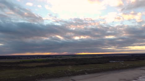 toma aérea de un hermoso horizonte de puesta de sol naranja nublado, rastreando hacia atrás a través de una playa y en lo profundo del océano, horsey gap, norfolk, inglaterra