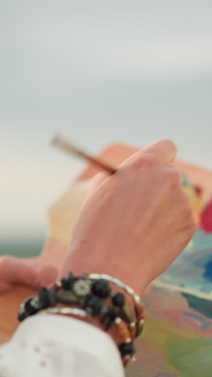 un primer plano de la mano de una mujer, adornada con cuentas, mezclando suavemente colores en una paleta con un pincel. la escena captura el delicado movimiento de mezcla de pintura bajo la suave luz del sol