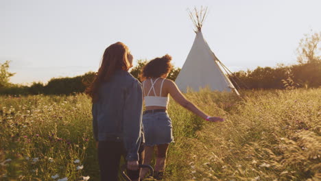 Dos-Amigas-Caminando-Tirando-Del-Carrito-A-Través-Del-Campo-Hacia-Un-Tipi-En-Vacaciones-De-Verano-En-Camping