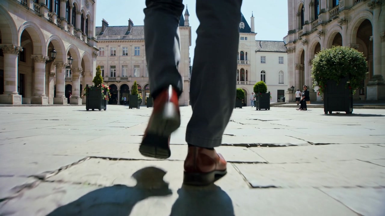 Left foot landing, man's feet walking across flagstone plaza to buildings, grey pants, brown shoes