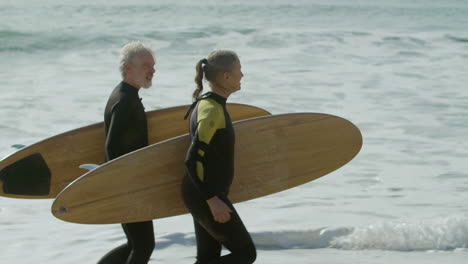 senior couple in wetsuit holding surfboard and running into the ocean