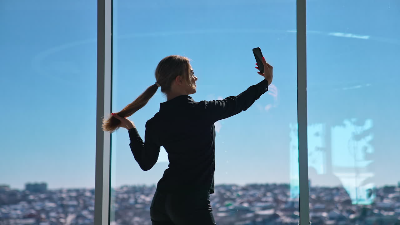 Lovely business woman in black taking photo of herself. Beautiful young woman doing selfie on smartphone while standing at office window