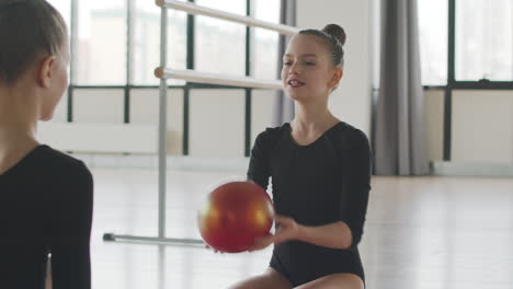 dos chicas rubias gimnásticas hablando mientras juegan con una pelota sentada en el suelo antes de comenzar la clase de ballet 2