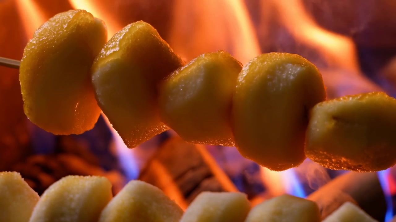 Vista ravvicinata di pezzi di patate pelate infilzati su uno spiedino di legno sopra braci e fiamme, che mostrano calore, doratura, superficie lucida, cucina rustica all'aperto, fumo e un ricco contrasto di colori.