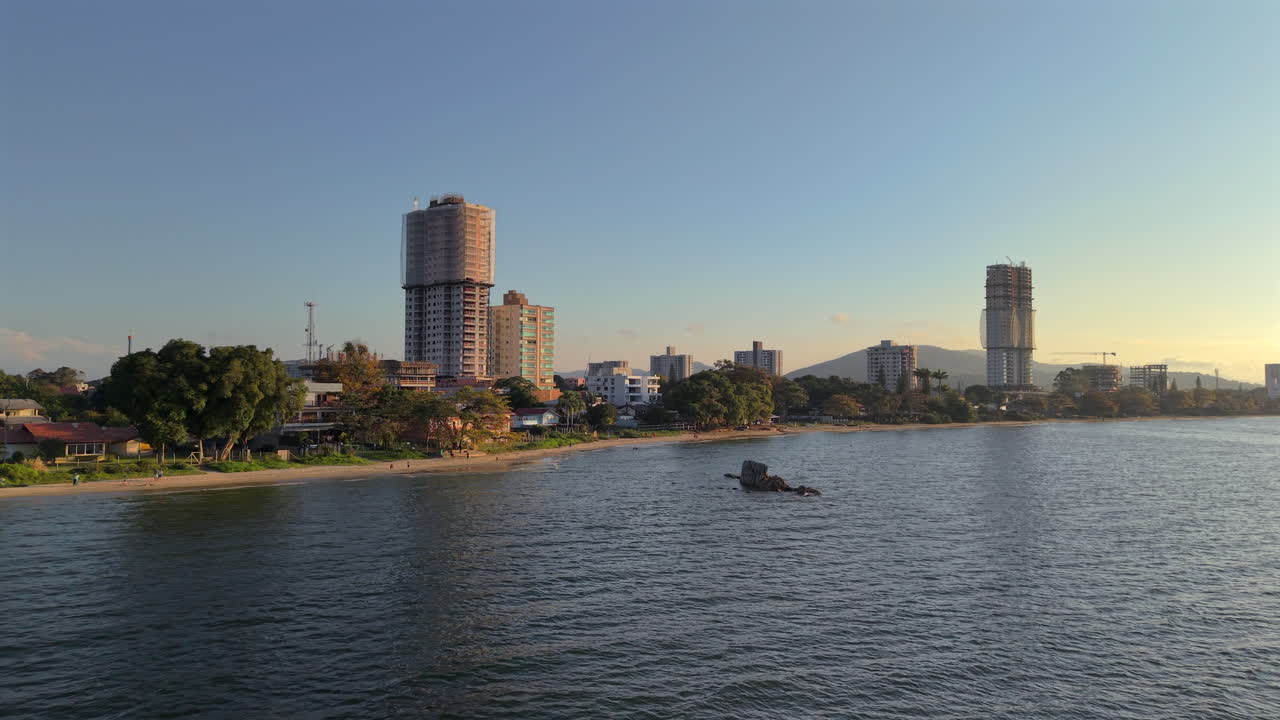 Aerial trucking shot flying along urban coastline with sandy beach of Penha, Santa Catarina, Brazil, revealing city skyline with new buildings under construction during a beautiful golden sunset