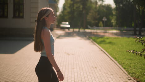 chic walking towards grassy area looking focused, wearing casual athletic wear, background featuring brick building, lush greenery, and blurred view of car near building, under warm sunlight