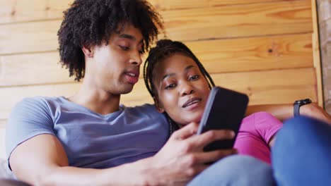 Happy-african-american-couple-using-smartphone,-sitting-on-sofa-in-log-cabin,-slow-motion