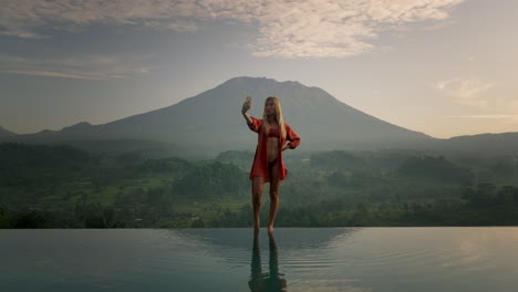 blond model posing on infinity pool edge in bikini during sunrise, bali mount agung