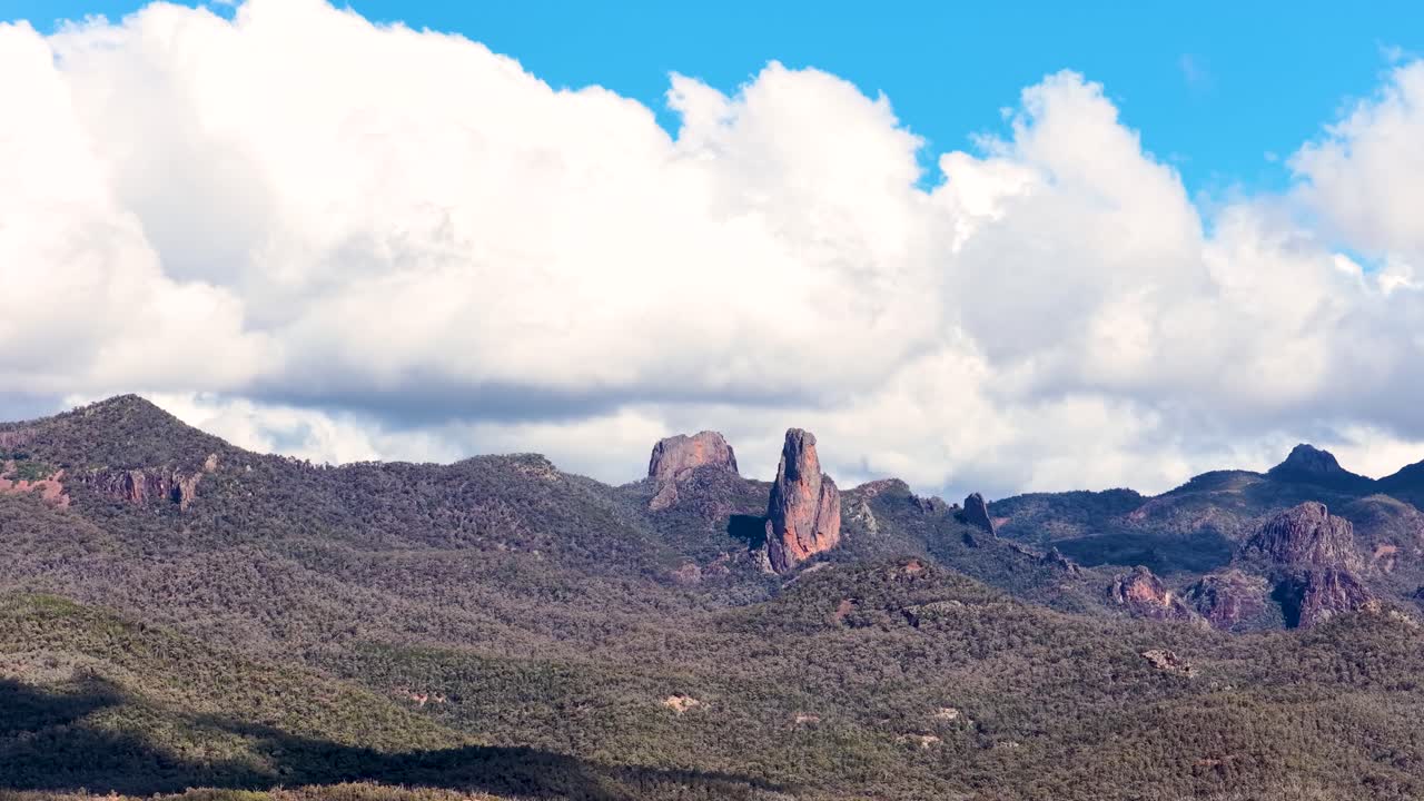 Camera pans across rugged Warrumbungle National Park mountains in daylight, revealing dramatic rock formations and shifting clouds under bright, natural lighting