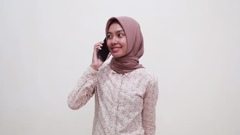 Young-asian-muslim-woman-standing-while-talking-on-phone-against-white-wall-background