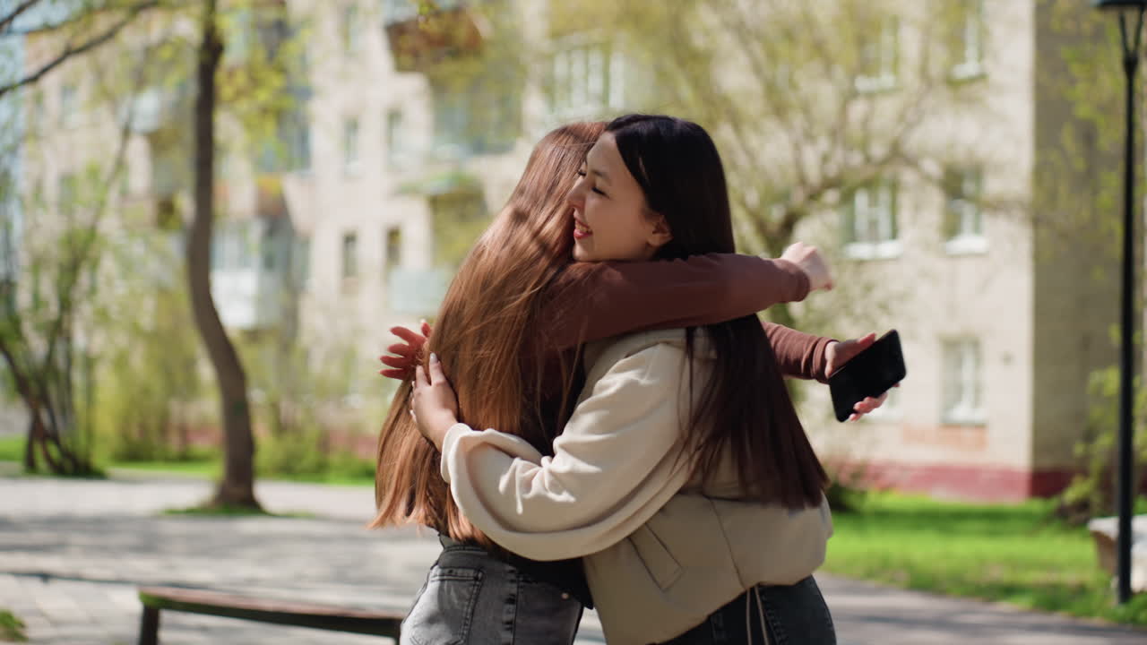 Mujer caucásica concentrada en el teléfono mientras su amiga asiática se acerca sigilosamente para abrazarla por detrás, una sonrisa rápida de sorpresa y luego un cálido abrazo, escena en un parque urbano con árboles, bancos y una suave luz primaveral