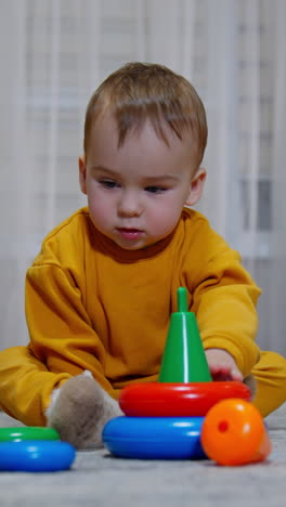 mom's hand takes a piece of toy pyramid and gives it to her kid. baby boy takes it, throws on the floor and picks another color ring leaving with it. vertical video