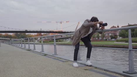 joven elegante con una cámara profesional tratando de encontrar una buena posición para una toma en una calle a lo largo de un río en una zona urbana