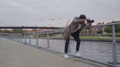 stylish young man with a professional camera trying to find a good position for a shot on a street along a river in an urban area