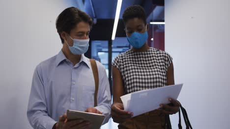 Diverse-business-colleagues-wearing-face-masks-using-tablet-in-discussion-walking-through-office