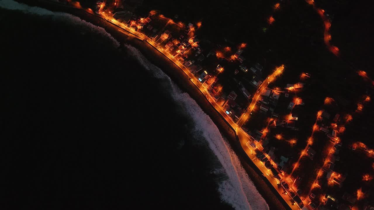 Admire the beauty of Madeira as the sun sets, with dramatic waves crashing on the coastline and the city lights twinkling along the water's edge, all captured from a breathtaking drone perspective