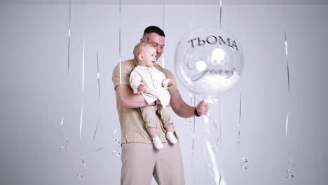 happy caucasian father with baby in hands in studio. lovely toddler plays with a big transparent balloon. white backdrop.