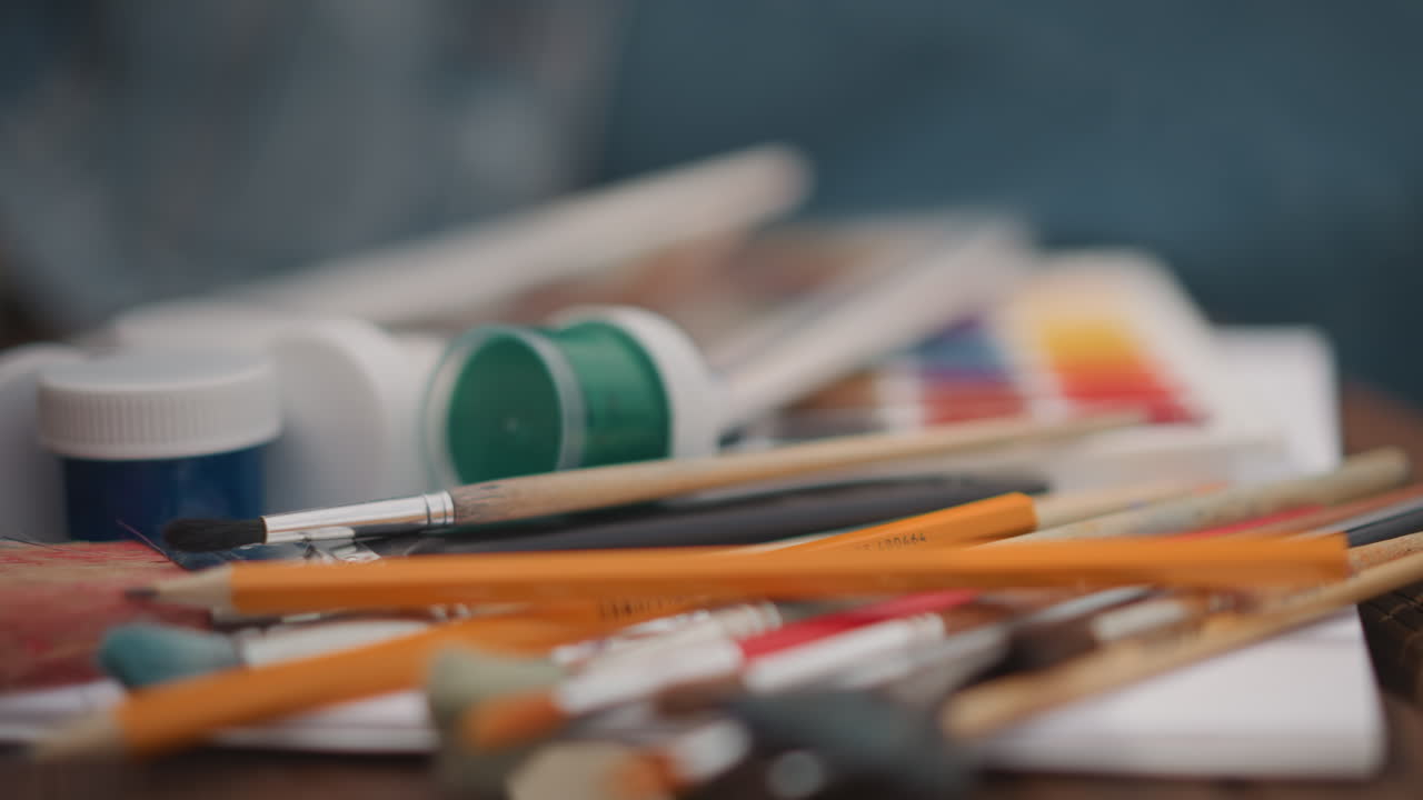 Table remplie de fournitures d'art et de pinceaux, crayons éparpillés, tubes et pots de peinture, pinceaux texturés, doigts tendant la main pour choisir un outil, atmosphère de studio chaleureuse avec une énergie créative désordonnée.
