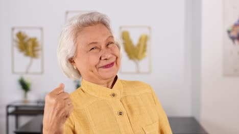 asian senior woman in yellow shirt smiles and makes a positive yes gesture in a well-lit, modern living room with soft natural lighting