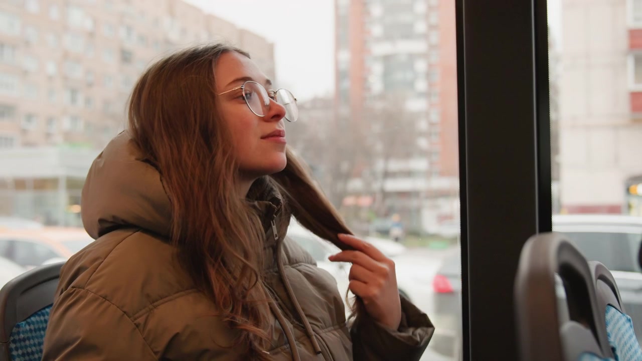 Woman on bus. Casual woman in urban transit scene. Young woman adjusts beanie amidst city apartment view. Relaxed female passenger wears puffer coat and glasses while observing city outside
