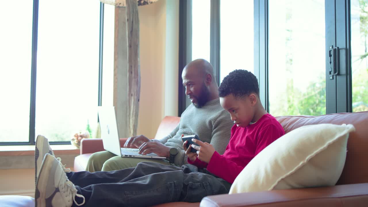 African American father and son bonding on sofa as father glancing at gaming device, typing laptop
