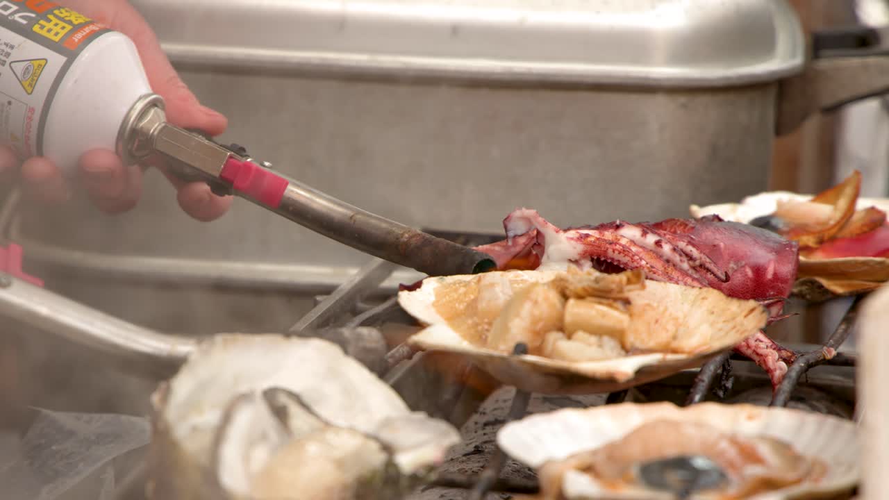 A close-up shot shows a vendor using a blowtorch to sear scallops and oysters