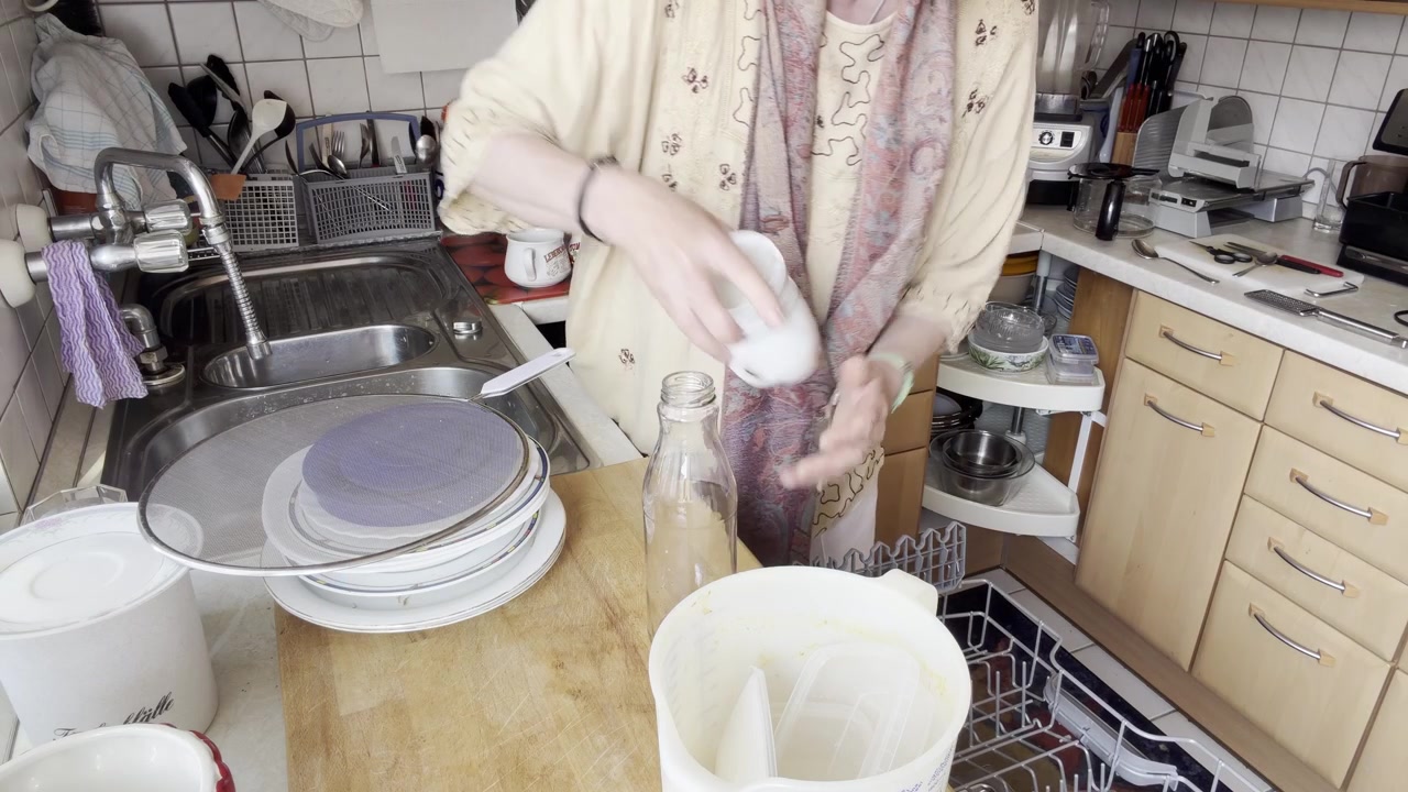 A static kitchen shot showing a woman loading cups into the upper dishwasher rack while removing a bottle to create more space for organized placement.