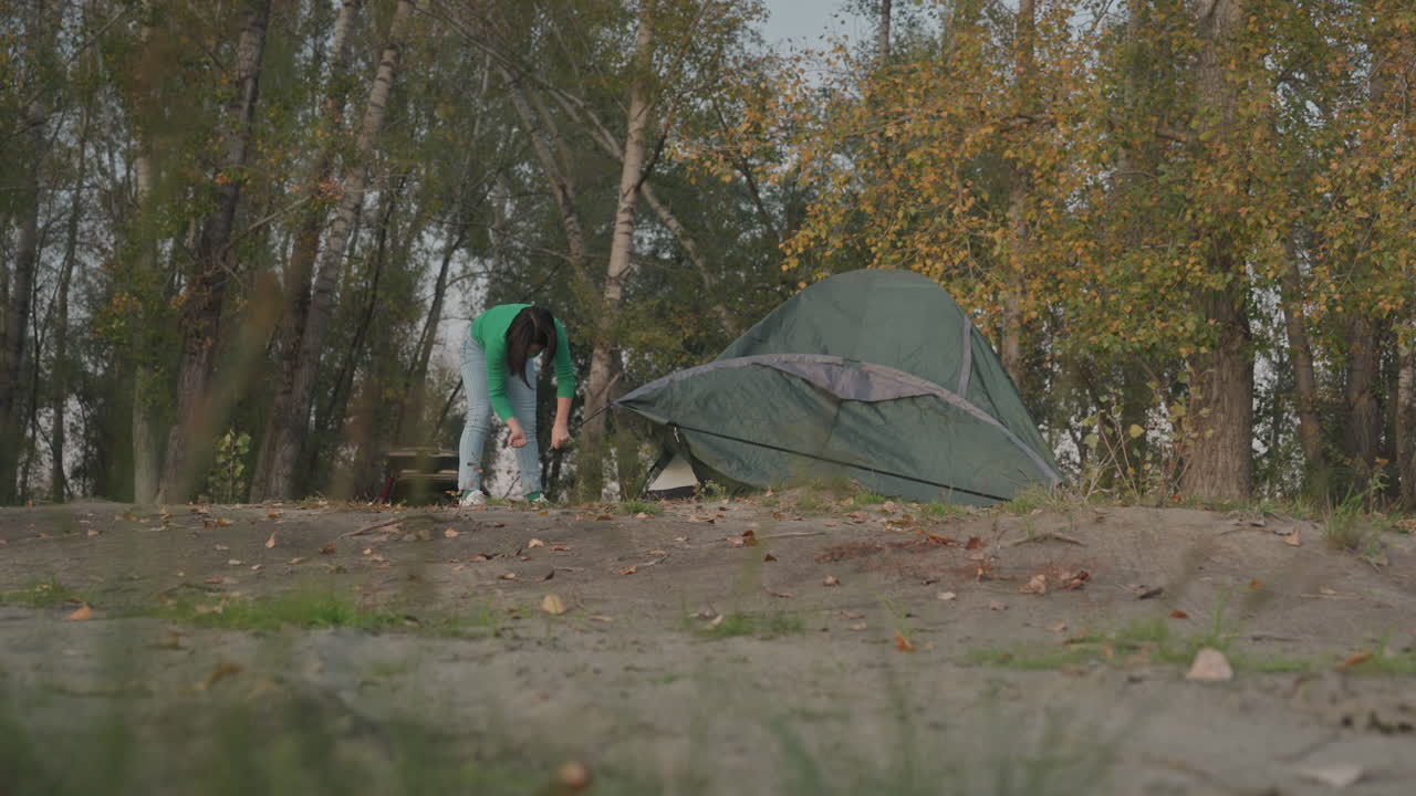 Person pakker telt sammen efter camping, folder stof og pakker stænger i taske, sikrer stropper og tjekker pløkker, blødt skumringslys, birketræer og lysning, stille afgangsstemning vises i billederne.