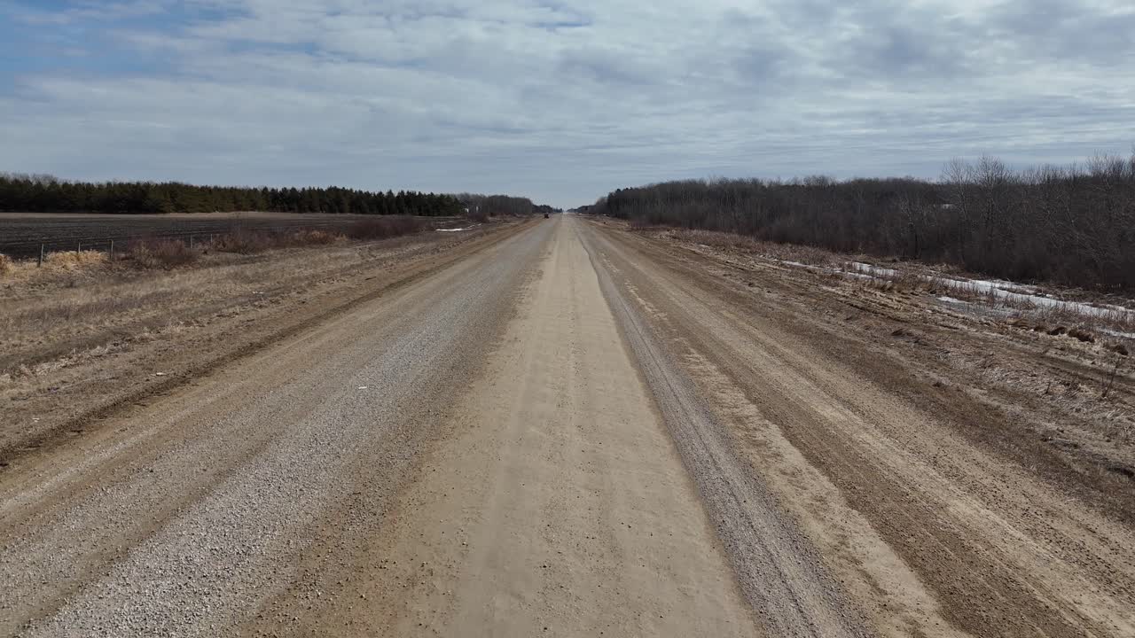 Låg drönarbild som följer en grusväg i södra Manitoba under våren, visar platt prärielandskap, bruna fält, glesa träd och öppet landsbygdslandskap under en klar himmel.