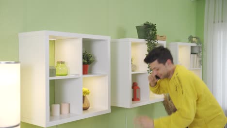 um jovem desajeitado a falar ao telefone em casa deixa cair um vaso de flores no chão.