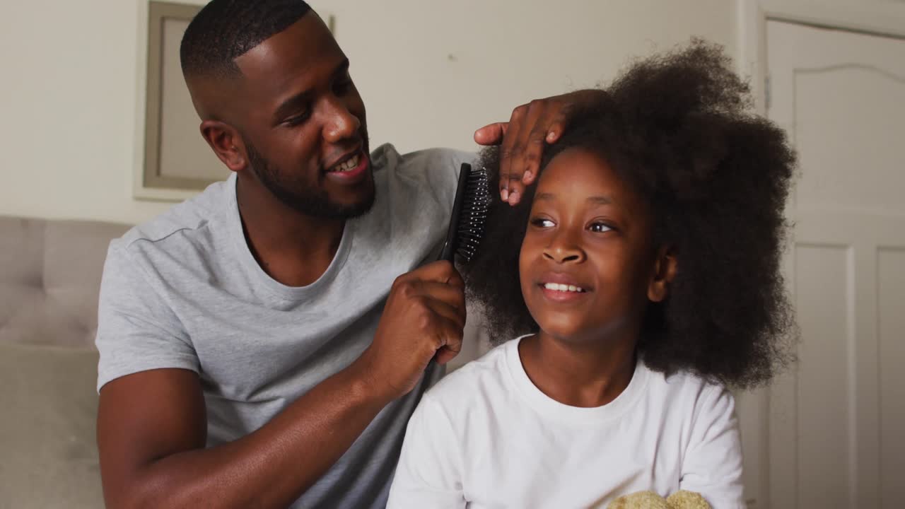 African american father brushing his daughters hair while sitting on the bed at home