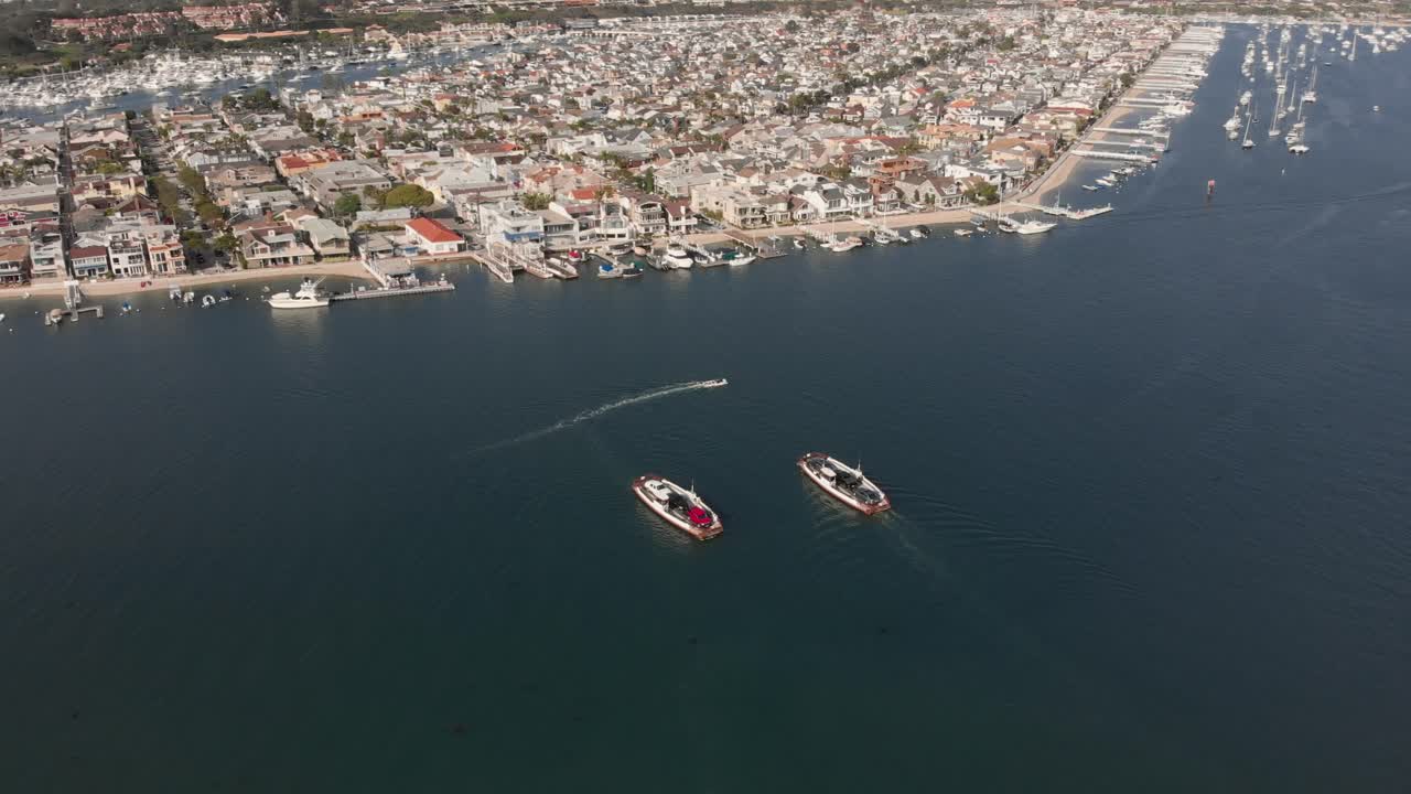 toma aérea orbitando alrededor de dos transbordadores que cruzan caminos en el puerto de newport con barcos anclados y edificios en el lado de la bahía y la isla balboa en el fondo en un día soleado de verano en el condado de orange