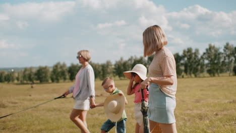 woman holding dog leash while boy carries sun hat and girl helps mother with picnic basket, walking across open field under blue sky with scattered clouds, surrounded by lush greenery and hay bale