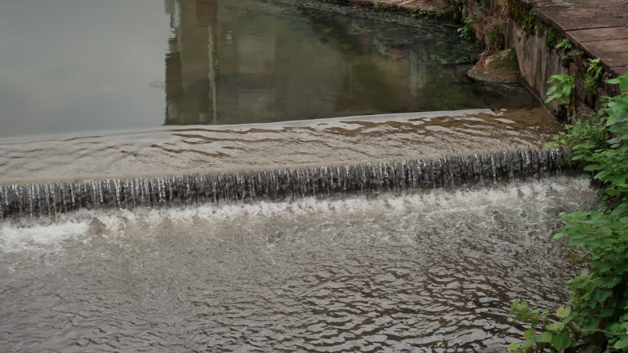 Piccolo sbarramento in cemento con acqua che scorre e riflessi nuvolosi sulla superficie, una dolce cascata che forma increspature e schiuma, vegetazione verde lussureggiante e pietre muschiose lungo la riva, corrente costante e movimento sottile