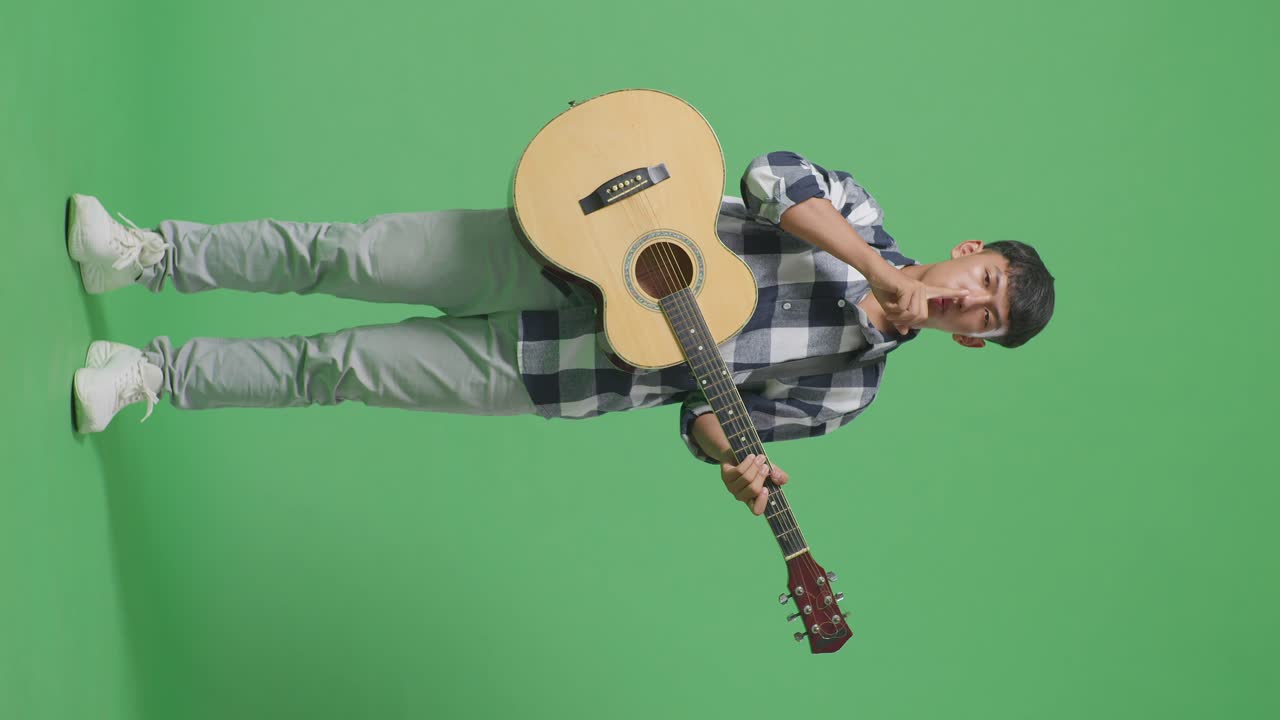 Full Body Of Asian Teenager With Guitar Showing Silence Gesture While Standing On Green Screen Background