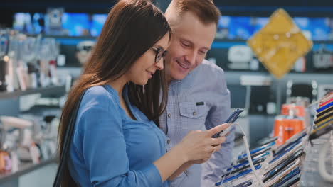 Young-beautiful-couple.-Man-and-woman-hold-in-hands-a-smartphone-standing-near-a-showcase-with-smartphones-in-the-appliances-store-choosing-the-best-smart-phone.-A-gift-to-his-wife