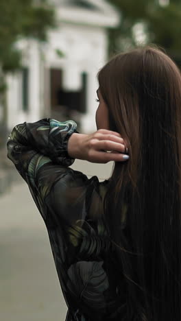 elegant young woman with long loose brown hair in stylish dress with floral print walks along city promenade slow motion close backside view