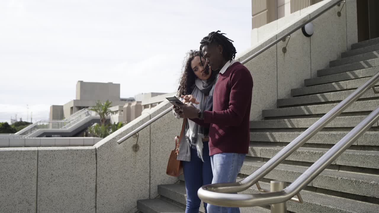 Stepping off staircase, Diverse friends pulling tablet, tapping smartphone for quick project review