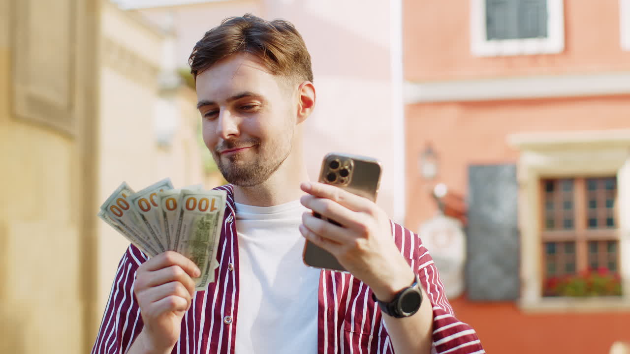 Happy bearded man counting money dollar cash use smartphone calculator app in urban city street