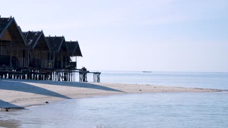 Row-of-wooden-beach-huts-overlooking-ocean-with-a-fishing-boat-traveling-by-on-a-tropical-island-in-Raja-Ampat,-West-Papua,-Indonesia