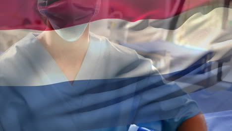 netherlands flag waving against caucasian female health worker wearing face mask at hospital