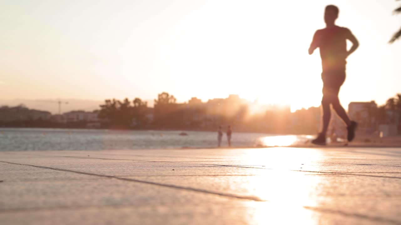 Slow motion, silhouette of man jogging on pavement by ocean during golden hour