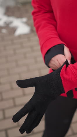 partial view of woman in red hoodie standing on interlocked path with snow along edges, putting on black winter gloves for warmth during cold weather in outdoor urban setting