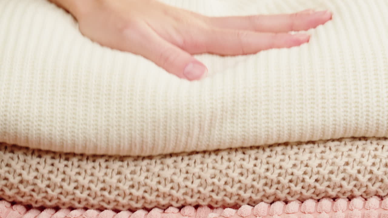 Woman's hand holding a stack of knitted sweaters in cream, beige, and pink