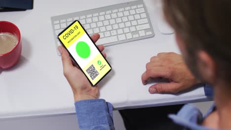 Man-at-desk-holding-smartphone-with-covid-vaccination-certificate-and-qr-code-on-screen